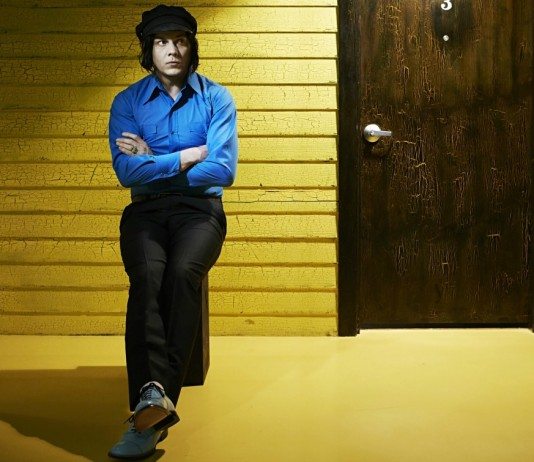 Jack White con camisa azul sobre un taburete delante de un muro amarillo.